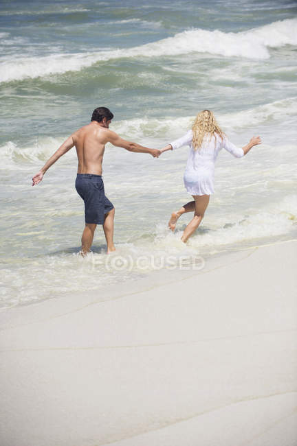Cheerful couple running on beach in wavy sea — Stock Photo