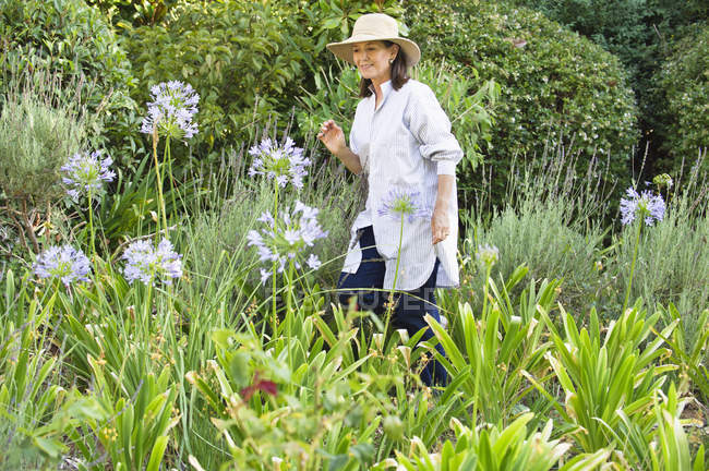 Mulher sorridente em chapéu de palha olhando para flores em um jardim — Fotografia de Stock