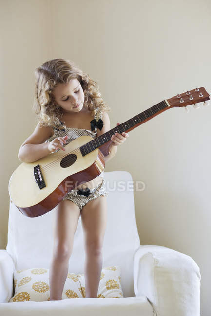 Cute little girl standing on armchair and playing a guitar — Stock Photo