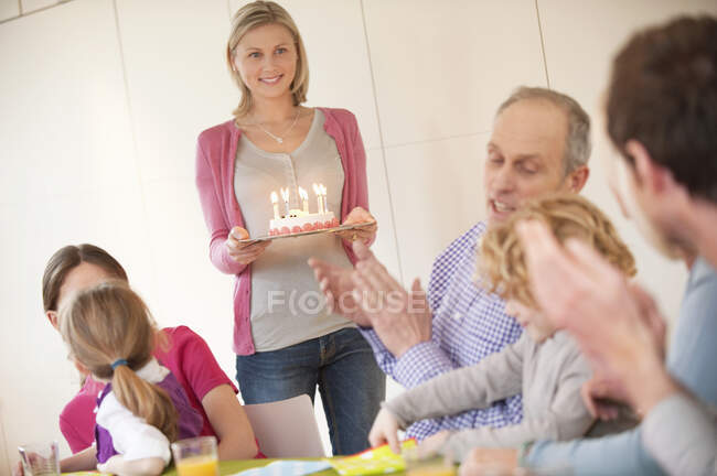 Familia en una celebración de cumpleaños con una mujer trayendo pastel en el fondo - foto de stock