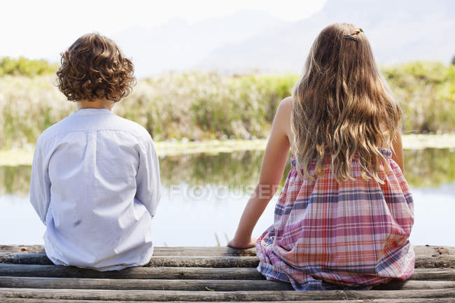 Вид на братьев и сестер, сидящих вместе за деревянным пиром на озере на природе — стоковое фото