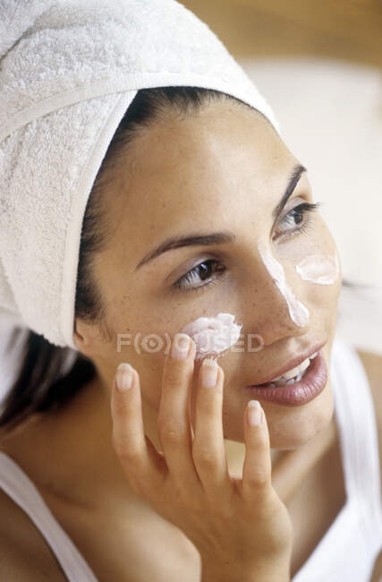 Primer plano de una joven con turbante de toalla, aplicándose crema en la cara - foto de stock