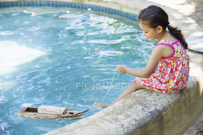 Menina sentada na borda da piscina e brincando com barco de brinquedo na água — Fotografia de Stock