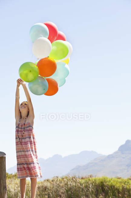 Menina em vestido xadrez brincando com balões coloridos no cais na natureza — Fotografia de Stock