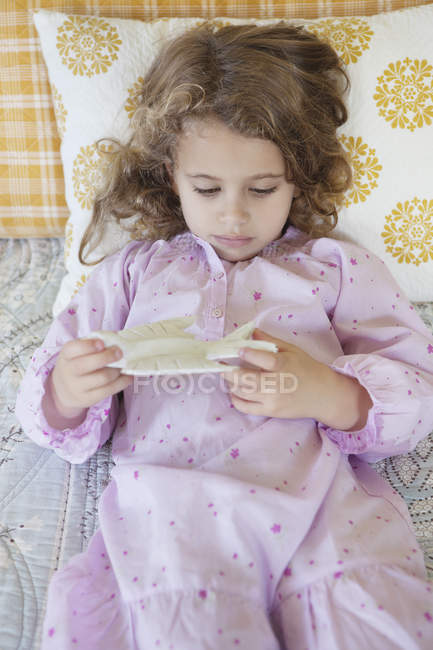 Милая маленькая девочка, играющая с игрушечной птичкой, пока лежит на кровати — стоковое фото