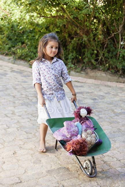 Nettes Mädchen schiebt Schubkarre mit Spielzeug in Garten — Stockfoto