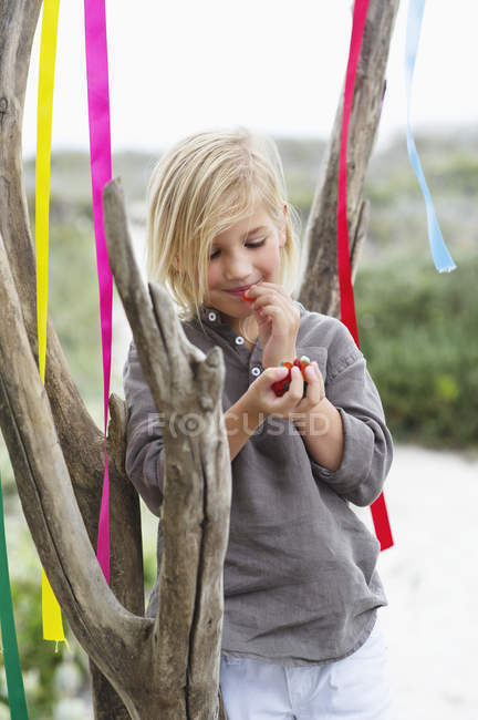 Girl leaning against tree with colorful ribbons and eating fruit — Stock Photo