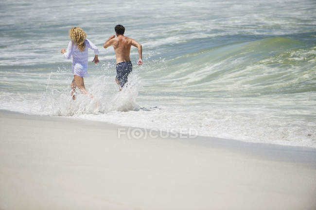 Rückansicht eines fröhlichen Pärchens, das am Strand im Wasser läuft — Stockfoto