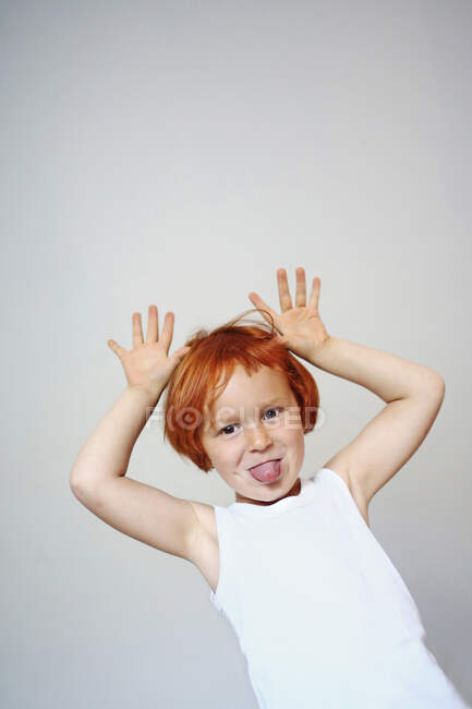 Retrato de una niña pelirroja en el interior sacando la lengua y levantando las manos - foto de stock
