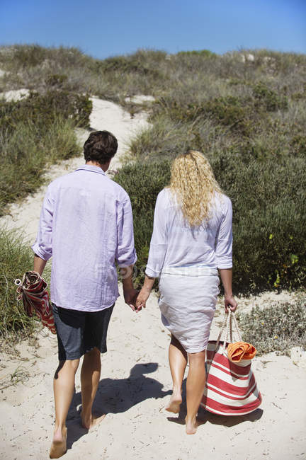 Вид сзади на пару, идущую по пляжу держась за руки с сумкой и пляжным зонтиком — стоковое фото