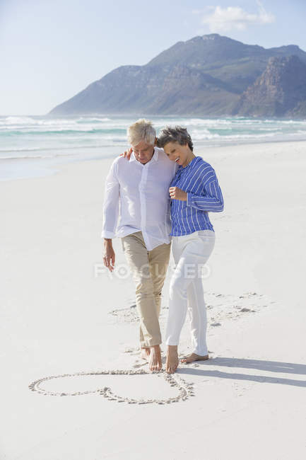 Abraçando casal sênior desenho forma de coração na areia com os pés — Fotografia de Stock