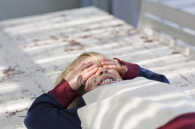 Playful boy lying on table outdoors with hands covering eyes — Stock Photo