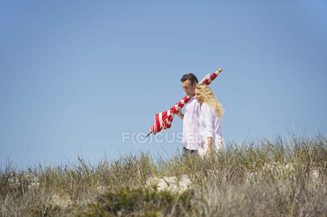 Пара, що йде на пляжі з смугастою пляжною парасолькою — стокове фото