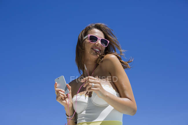 Donna felice in costume da bagno ascoltando musica con il cellulare di fronte al cielo blu — Foto stock