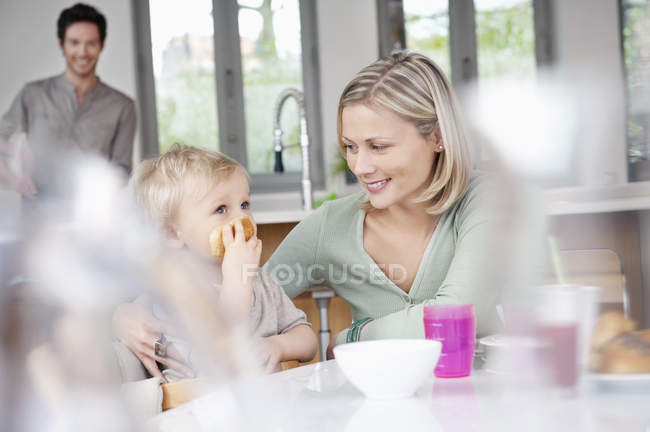Donna che fa colazione con piccolo figlio in cucina con marito sullo sfondo — Foto stock