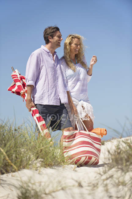 Feliz pareja caminando en la playa con bolsa y sombrilla de playa - foto de stock