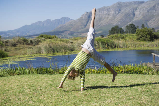 Cute little boy doing cartwheel on grass in nature against mountains — Stock Photo