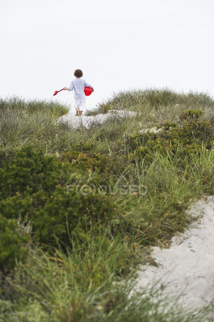 Boy running on sand dunes on beach with red ball and shovel — Stock Photo