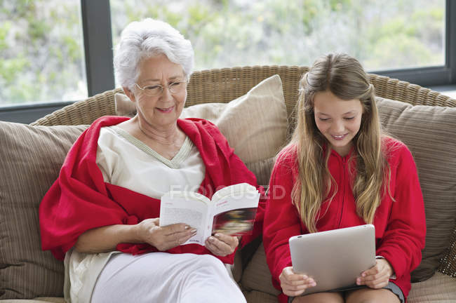 Mujer mayor leyendo libro con nieta usando tableta digital - foto de stock