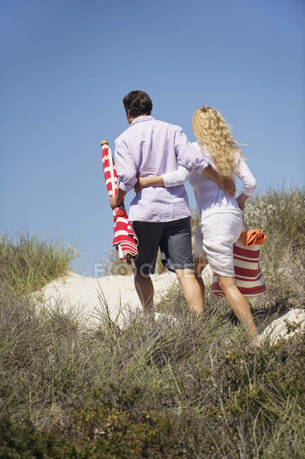 Vue arrière du couple embrassant marchant sur la plage avec sac et parasol — Photo de stock