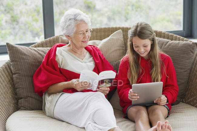 Mujer mayor leyendo libro con nieta usando tableta digital - foto de stock