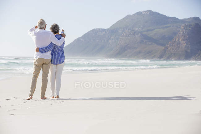 Vista trasera de abrazar a la pareja de pie en la playa y mirando a la vista - foto de stock