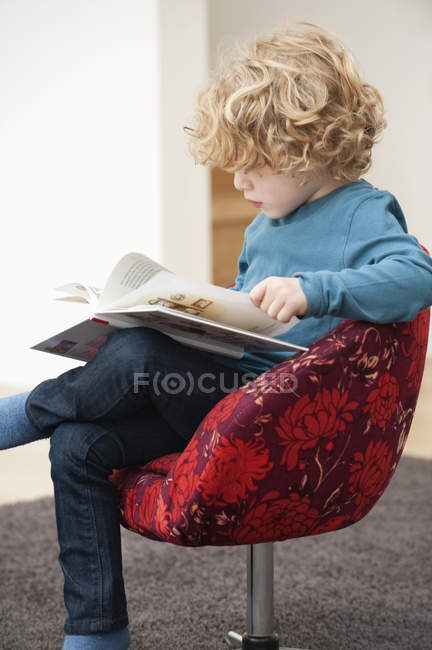 Menino bonito com cabelo loiro lendo um livro em poltrona em casa — Fotografia de Stock