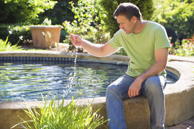 Homem maduro sentado à beira da piscina no jardim e tocando água — Fotografia de Stock