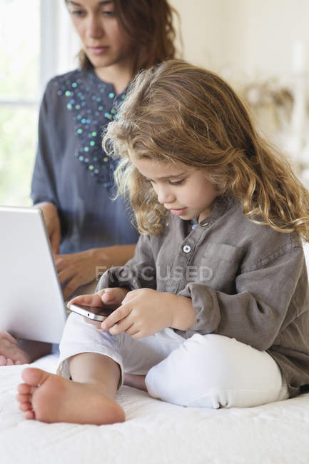 Девушка смотрит на смартфон с матерью, работающей на ноутбуке на кровати — стоковое фото