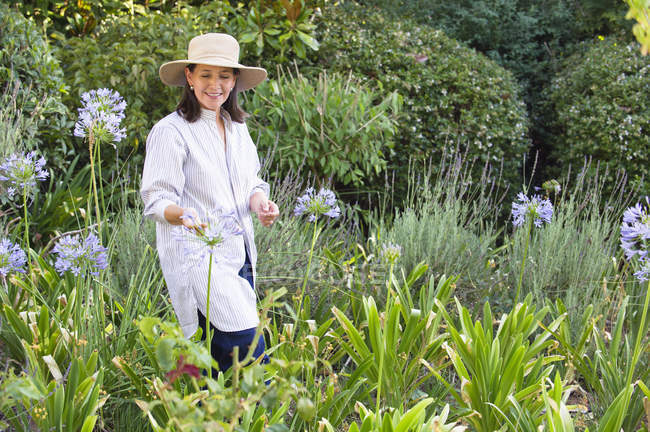 Donna sorridente in cappello di paglia che guarda fiori in un giardino — Foto stock