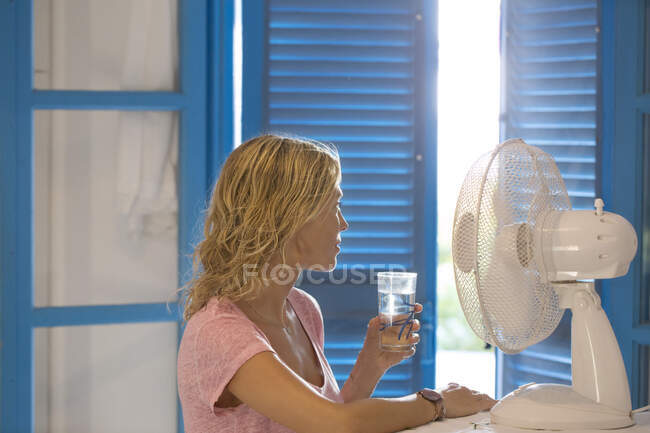 Giovane donna di profilo di fronte a un ventilatore con un bicchiere d'acqua. — Foto stock
