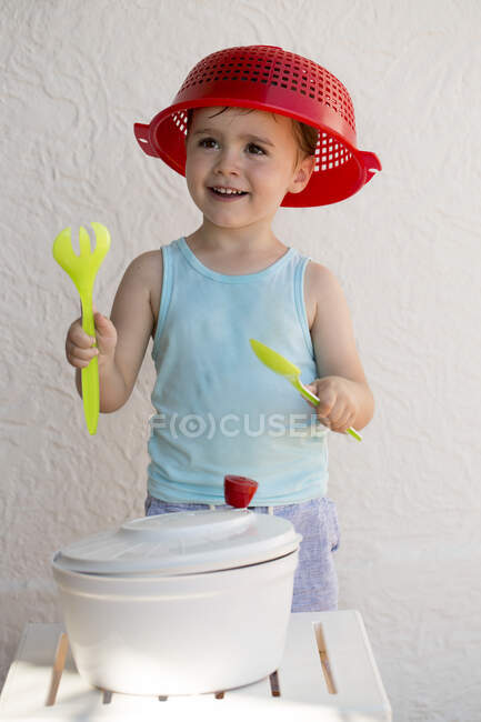 Niño con un escurridor en la cabeza divirtiéndose con un escurridor y cubiertos de ensalada. - foto de stock