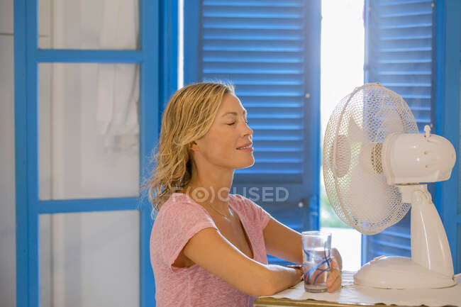 Junge Frau im Profil vor einem Ventilator mit einem Glas Wasser. — Stockfoto