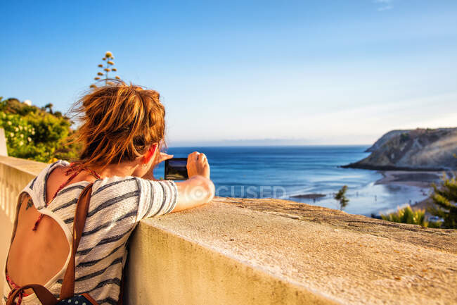 Молодая женщина сзади, фотографируется со своим смартфоном на низкой стене с пляжа Бургау, Алгарве, Португалия — стоковое фото