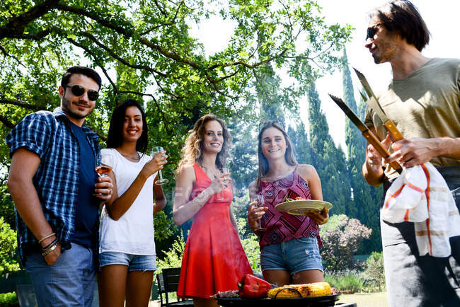 Grupo de jóvenes felices y alegres divirtiéndose alrededor de la parrilla de barbacoa durante las vacaciones de verano fiesta al aire libre en el jardín. - foto de stock