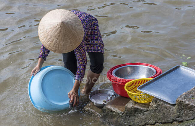 Vietnam, Delta del Mekong, Can Tho, mujer con un sombrero cónico asiático lavando sus platos en el río - foto de stock