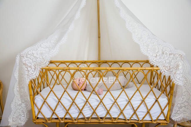 View of a baby awake 2 months in beige layette lying on his back in his baby bed canopy wicker and lace. — Stock Photo