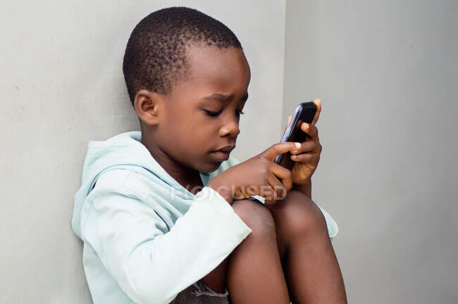 Niño sentado contra la pared manipulando un teléfono móvil con curiosidad. - foto de stock