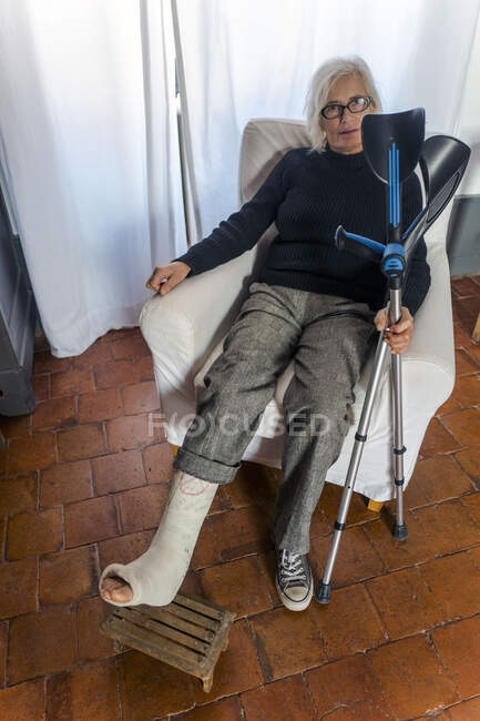 Femme assise sur une chaise avec une jambe plâtrée sur un repose-pieds et tenant ses béquilles — Photo de stock