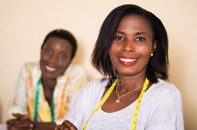 Smiling seamstress in her workshop looks at the camera with her colleague in the background. — Stock Photo