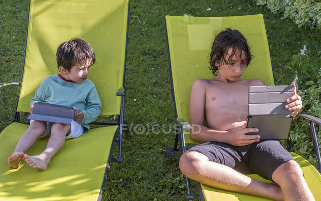 Dos chicos de cuatro años y doce años, sentados en tumbonas con su tablet digital - foto de stock
