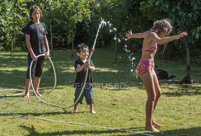Два мальчика 4 года и 12 лет и 10-летняя девочка, играющая со шлангом в саду — стоковое фото