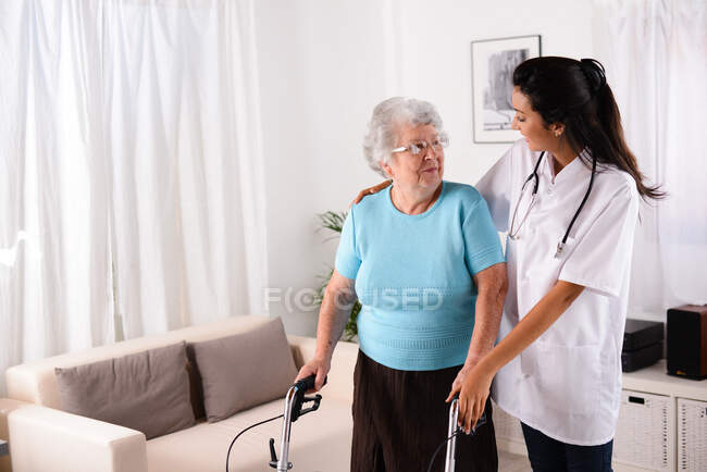 Весела молода медсестра реабілітації, яка допомагає літній старшій жінці, використовуючи ходунки — стокове фото