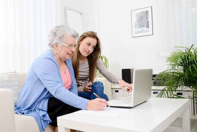 Joven alegre ayudando a una mujer mayor en casa con el ordenador portátil - foto de stock