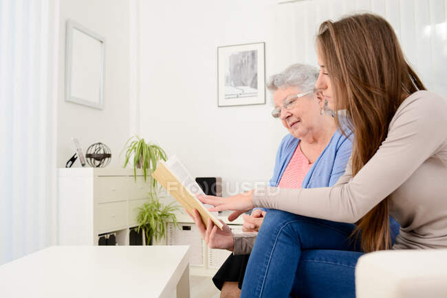 Cheerful young woman reading book for old senior woman at home — Stock Photo