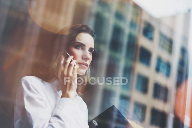 Photo business woman wearing modern white shirt, talking smartphone and holding documents in hands. Open space loft office. Panoramic windows background. Horizontal mockup. Film effect — Stock Photo