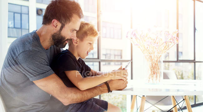 Niño sentado con el padre en la mesa y jugando juntos pc tableta en el loft moderno. Fondo horizontal, borroso. Efecto luces solares . - foto de stock