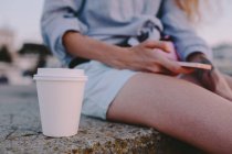 Біла пластикова чашка кави біля сидячої жінки зі смартфоном — стокове фото
