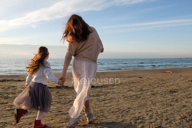 Вид сзади матери с дочерью, держащейся за руки во время прогулки по пляжу — стоковое фото