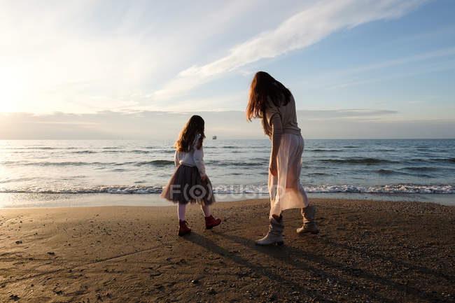 Vue arrière de la mère avec sa fille debout à la plage contre le ciel avec des nuages — Photo de stock